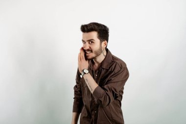 Kahverengi gömlekli genç adam beyaz arka planda yalnız poz veriyor. Ağız üstünde el ele, gizli dedikodular, kötü niyetli konuşmalar..