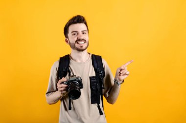 Sarı arka planda izole edilmiş bej tişört giyen mutlu genç adam kameraya bakıyor ve profesyonel bir kamera tutarken işaret parmağıyla işaret ediyor..