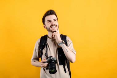 Sarı arka planda izole edilmiş bej tişört giyen, kendinden emin gülümseyen, boş görünüşlü, gülümseyen ve elinde bir dslr kamerayla çenesine dokunan genç bir adam..