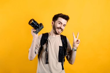 Mutlu genç adam bej tişört giyiyor. Sarı arka planda yalnız poz veriyor. Elinde DSR kamerayla gülümsüyor..