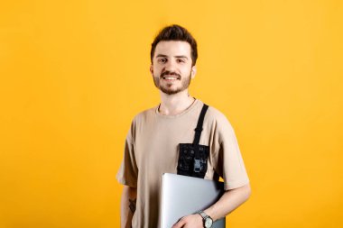 Beyaz tişört giyen, sarı arka planda yalnız poz veren, dizüstü bilgisayarda poz veren beyaz bir adam. Gülümseyerek kameraya bakıyorsun..