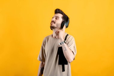 Sarı arka plan kablosuz kulaklıklar üzerine izole edilmiş bej tişört giyen genç adam meditasyon yapıyor. Kulaklığa dokunuyorum..