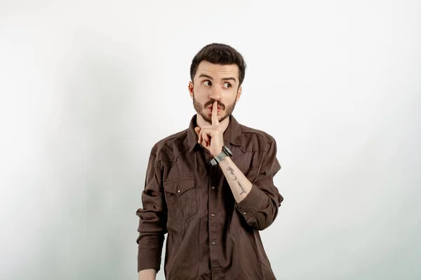 Handsome caucasian man wearing casual clothes posing isolated over white background makes shush gesture, looks surprisingly aside, notices something strange, has dark stubble.