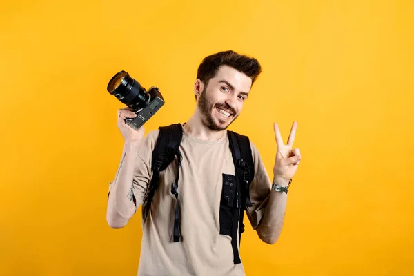 Happy young man wearing beige t-shirt posing isolated over yellow background man holding dslr camera and smiling with happy face looking at the camera doing victory sign with fingers.