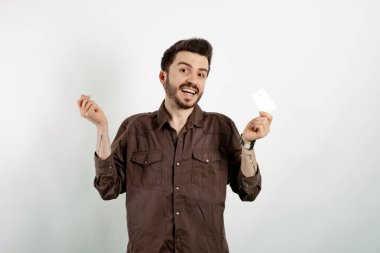 Kahverengi gömlekli neşeli beyaz adam beyaz arka planda soyutlanmış poz veriyor. Elinde kredi kartı ve para işareti, gülen surat ve mutlu kazançlar var.