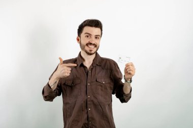 Kahverengi gömlekli neşeli bir genç beyaz arka planda izole edilmiş bir şekilde poz veriyor. Kredi kartı ya da banka kartını işaret ediyor. Alışveriş ve finans kavramı.