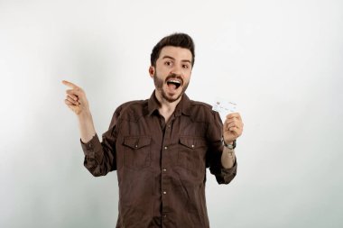 Beyaz arka planda yalnız poz veren beyaz gömlekli genç beyaz bir adamın portresi. Elinde kredi kartı ve işaret parmağı var. Alışveriş ve finans kavramı.