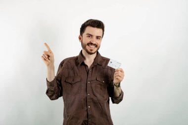 Kahverengi gömlekli genç adam beyaz arka planda yalnız poz veriyor. Elinde kredi kartı var ve elini ve parmağını kenarı işaret ediyor..