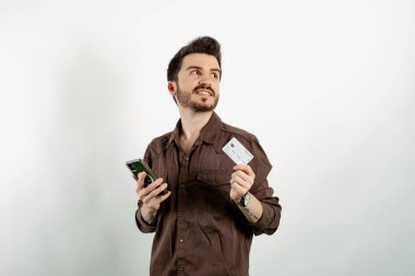 Beyaz arka planda yalnız poz veren beyaz tişörtlü genç bir adam kredi kartıyla cep telefonuyla alışveriş yapıyor. Online alışveriş kavramları.