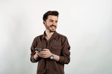 Yakışıklı, sıradan kıyafetler giyen, beyaz arka planda soyutlanmış, hayalperest görünümlü, akıllı telefonu tutarken düşünen ve yan bakan genç bir adam..