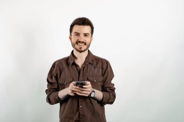 Kahverengi gömlekli genç adam beyaz arka planda yalnız poz veriyor akıllı bir telefon tutuyor ve kameraya büyük bir gülümsemeyle bakıyor. Sosyal medyada mesajlaşma, izleme veya kaydırma.