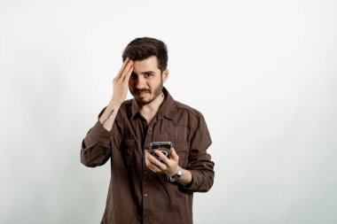 Kahverengi gömlekli genç adam beyaz arka planda yalnız poz veriyor telefonda konuşurken çaresiz hissediyor. Mobil konuşma sırasında kötü haberler duymak.