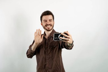 Mutlu, sıradan kıyafetler giyen, beyaz arka planda izole edilmiş bir şekilde poz veren, selfie çeken ya da video çekip el sallayan bir adam. Vlog kaydı, sosyal medya etkileyicisi yayınlanıyor, video çağrısı yapılıyor.