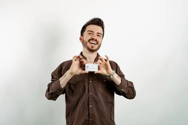 Happy young man wearing shirt posing isolated over white background showing his plastic credit or debit card with both hands. Shopping and finance concept.