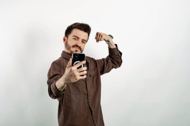 Beyaz arka planda yalnız poz veren beyaz gömlekli beyaz bir adam selfie çekiyor, güçlü kolunu gösteriyor, pazılarını esnetiyor ve fotoğraf için poz veriyor..