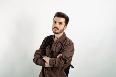 Beyaz arka planda yalnız poz veren beyaz tişörtlü genç bir adam sırt çantasında çapraz kollarla poz veriyor..
