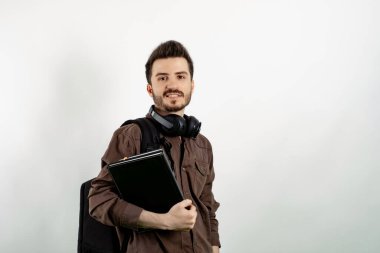 Sırt çantası ve kitaplarla izole edilmiş beyaz bir öğrenciye poz verip kameraya bakarak kendinden emin gülümseyen genç bir adam. Lise üniversite konsepti.