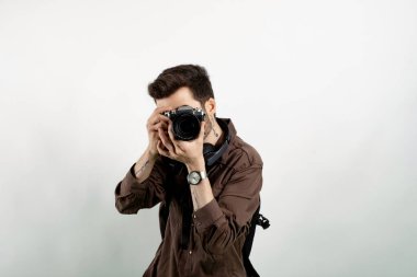 Yakışıklı, günlük kıyafetler giyen, beyaz arka planda yalnız poz veren ve DSR kamerasıyla fotoğraf çeken genç bir adam. Fotoğrafçı yüzünü kamerayla kapatıyor..