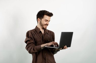 Yüzünde mutlu ve havalı bir gülümsemeyle dizüstü bilgisayar kullanarak beyaz arka planda izole edilmiş rahat kıyafetler giyen genç adam gülümsüyor. Dişler gösteriliyor.