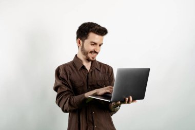Kahverengi gömlekli neşeli genç adam beyaz arka planda yalnız poz veriyor ve dizüstü bilgisayar kullanıyor yüzünde mutlu ve havalı bir gülümsemeyle gülümsüyor. Dişler gösteriliyor.