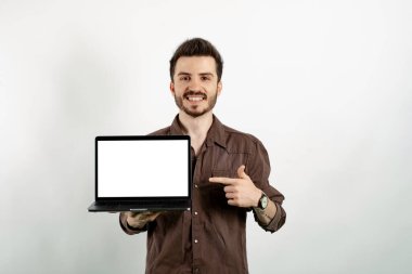 Kendine güvenen, rahat kıyafetler giyen, beyaz arka planda izole edilmiş bir şekilde duran genç bir adam işaret parmağıyla boş ekran ile dizüstü bilgisayara işaret ediyor.