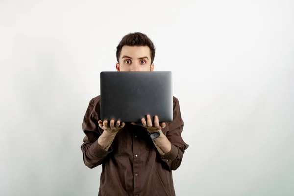 Young man wearing brown shirt posing isolated over white background peeking out from behind laptop, looking surprised at camera.