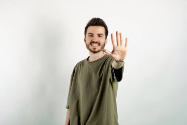 Yeşil tişört giyen, kendine güvenen genç adam beyaz arka planda izole edilmiş bir şekilde poz veriyor ve beş numaralı parmaklarıyla gülümseyerek işaret ediyor..