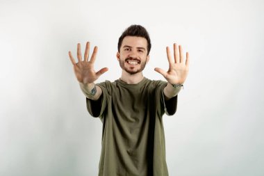 Yeşil tişört giyen, kendine güvenen genç adam beyaz arka planda izole edilmiş bir şekilde poz veriyor ve on numaralı parmaklarıyla gülümseyerek onu işaret ediyor..