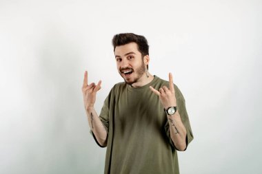 Yeşil tişört giyen mutlu genç adam beyaz arka planda izole edilmiş bir şekilde duruyor ve elleri yukarıda çılgın bir ifadeyle rock sembolü yapıyor. Müzik yıldızı. Ağır konsept.