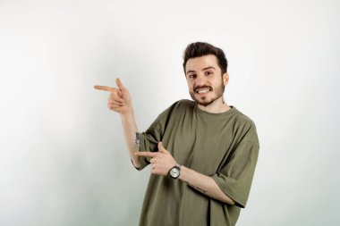 Neşeli beyaz bir adam beyaz arka planda soyutlanmış bir şekilde poz veriyor gülümsüyor ve kameraya bakıyor iki eliyle ve parmaklarıyla sol tarafı işaret ediyor..