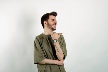 Beyaz arka plan üzerinde izole edilmiş yeşil tişört giyen yakışıklı beyaz adam başparmağıyla yana bakıyor ve işaret ediyor..