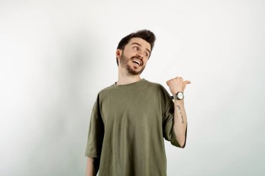 Yakışıklı, günlük kıyafetler giyen, beyaz arka planda izole edilmiş bir şekilde poz veren ve baş parmağıyla yan tarafı gösteren genç bir adam..