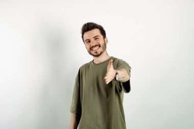 Kendinden emin gülümseyen genç bir adam beyaz arka planda izole bir şekilde poz veriyor kameraya uzanmış el sıkışma teklif ediyor. Dayanışma ve güven göstergesi.