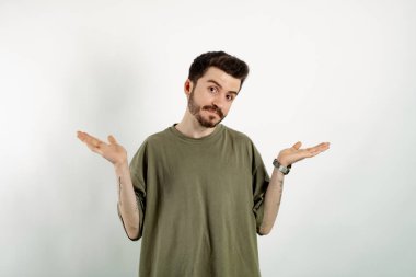 Beyaz arka planda yalnız poz veren beyaz tişörtlü yakışıklı beyaz adam, omuzlarını silkeliyor, şaşkın görünüyor, ellerini yana açıyor, bilemiyorum, belli olmuyor, duruyor.