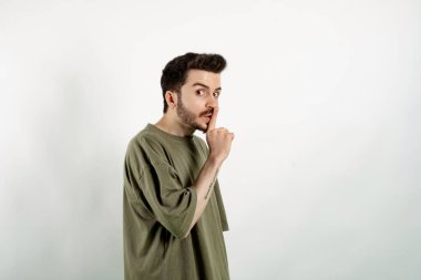 Beyaz arka planda yalnız poz veren beyaz tişört giyen beyaz bir adam şşşş hareketi yapıyor, şaşırtıcı bir şekilde yana, garip bir şey fark ediyor, siyah sakalı var..