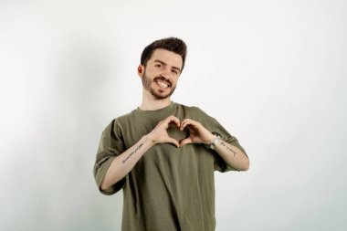 Kendinden emin gülümseyen genç bir adam beyaz arka planda izole bir şekilde poz veriyor ve parmaklarıyla kalp figürünü gösteriyor. Sevgiyle gülümseyen, elleriyle kalp sembolü yapan. Romantik konsept.