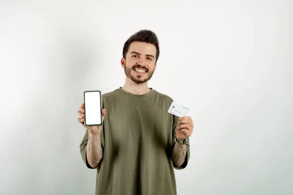 Beyaz arka plan boş ekran ve kredi kartı üzerinden izole edilmiş haki rengi tişört giyen genç adam, alışveriş veya çevrimiçi ödeme, bankacılık, modelleme için en yeni mobil uygulamayı tavsiye ediyor.