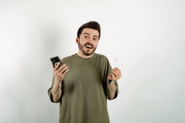 Beyaz arka plan üzerinde izole poz veren beyaz tişörtlü beyaz bir genç elinde kredi kartı veya banka kartı ve akıllı telefon tutuyordu..