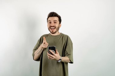 Kendinden emin gülümseyen genç bir adam beyaz arka planda izole edilmiş bir şekilde poz veriyor akıllı bir telefon tutuyor ve kameraya gülümsüyor. Sosyal medyada mesajlaşma, izleme veya kaydırma.