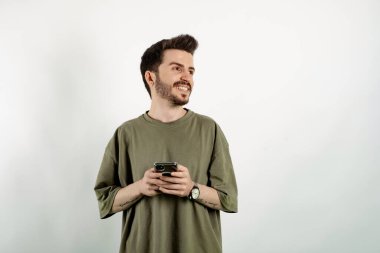 Haki tişört giyen genç yakışıklı bir adamın portresi beyaz arka planda yalnız poz veriyor, akıllı telefonu tutarken düşünüyor ve yan tarafa bakıyor..