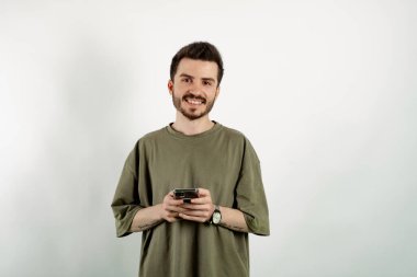 Haki tişört giyen genç yakışıklı bir adamın portresi beyaz arka planda izole bir şekilde poz veriyor akıllı bir telefon tutuyor ve kameraya gülümsüyor. Sosyal medyada mesajlaşma, izleme veya kaydırma.