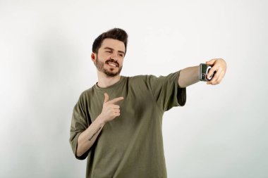 Haki tişört giyen genç adam beyaz arka planda izole edilmiş bir şekilde cep telefonuna işaret ediyor ve selfie çekiyor. İletişim kavramları.
