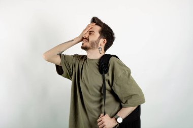 Beyaz arka planda izole edilmiş pozlar veren beyaz tişörtlü genç bir adam baş ağrısı çekiyor ve alnından el ele tutuşuyor. Üniversite eğitimi, sırt çantası konsepti.