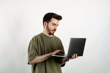 Haki tişört giyen genç adam beyaz arka plan üzerinde izole edilmiş bir şekilde duruyor ve dizüstü bilgisayar kullanıyor yüzünde mutlu ve havalı bir gülümsemeyle gülümsüyor. Dişler gösteriliyor.