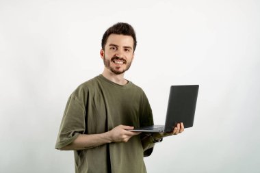 Beyaz arka planda izole edilmiş bir şekilde haki tişört giyen mutlu genç adam kameraya bakıyor ve yüzünde mutlu ve havalı bir gülümsemeyle gülümseyen bir bilgisayar dizüstü bilgisayar kullanıyor. Dişler gösteriliyor.
