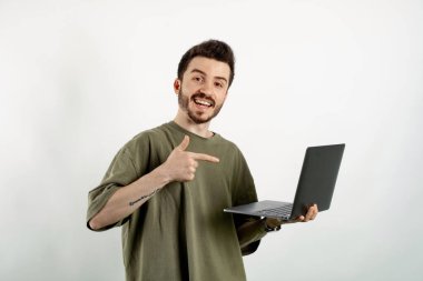 Kendinden emin gülümseyen genç adam beyaz arka planda izole edilmiş bir şekilde poz veriyor gülümsüyor ve işaret parmağıyla bilgisayarı işaret ediyor..