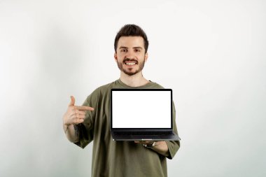 Kendinden emin gülümseyen genç adam beyaz arkaplan üzerinde yalnız poz vererek boş ekran ile dizüstü bilgisayara işaret ediyor.