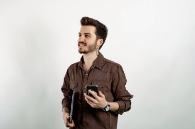 Kahverengi gömlekli neşeli beyaz bir adam beyaz arka planda yalnız poz veriyor dizüstü bilgisayar ve telefon pozu ile el ele tutuşuyor. Uzaklara bakıyorum..