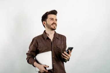 Sıradan kıyafetler giyen mutlu bir adam beyaz arka plan üzerinde soyutlanmış bir şekilde el ele tutuşup dizüstü bilgisayar ve telefon pozu veriyor. Uzaklara bakıyorum..