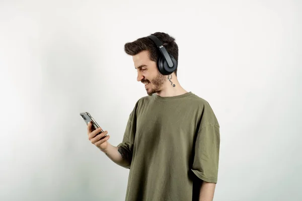Happy casual man wearing t-shirt posing isolated over white background wireless headphones listening to music from smartphone looking at the phone screen.